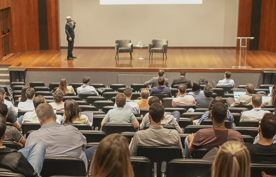 Foto da visão traseira de um auditório, com pessoas que assistem a uma palestra ao fundo