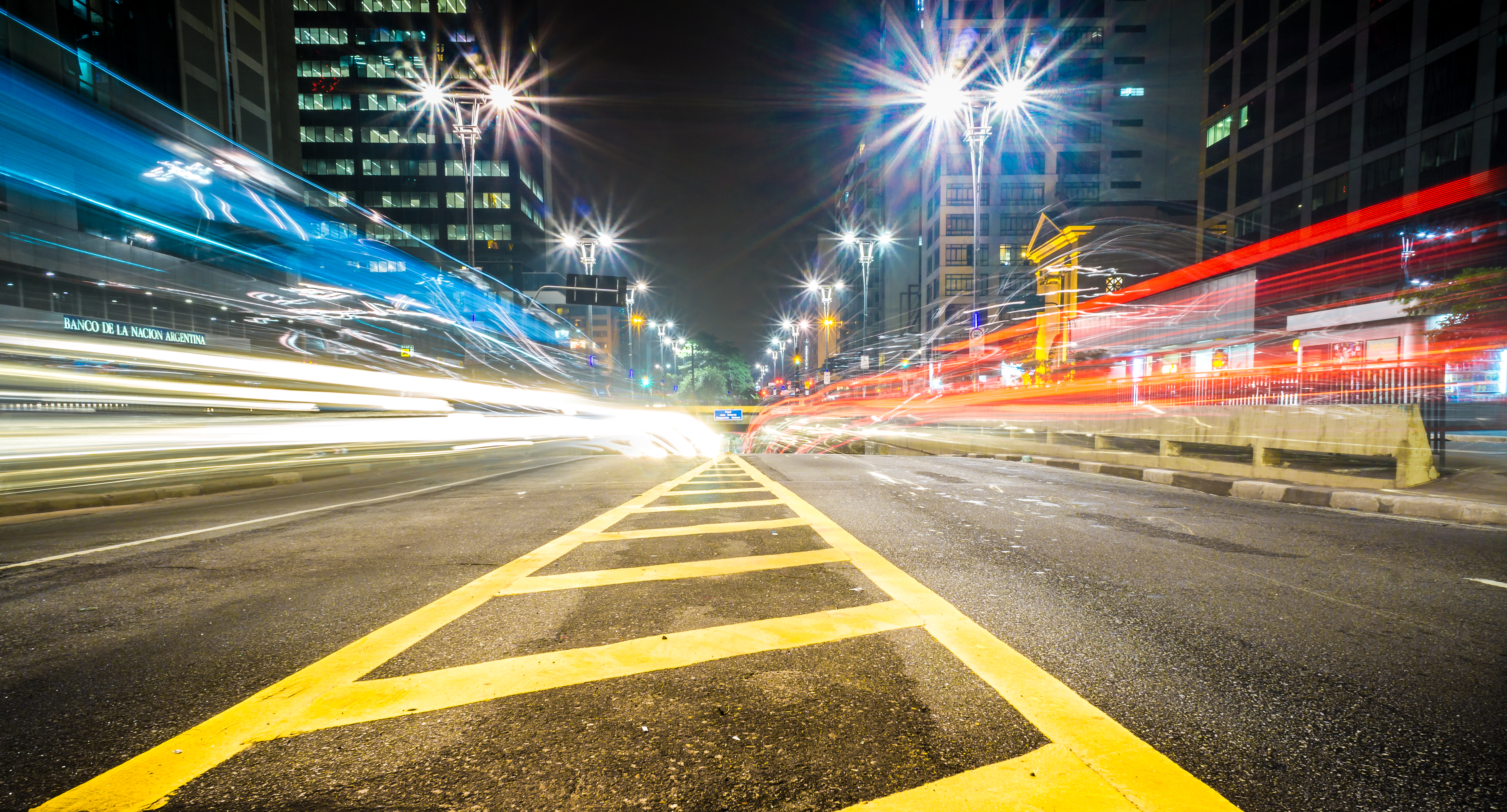 Imagem noturna da Avenida Paulista, com carros em movimento, prédios e iluminação pública