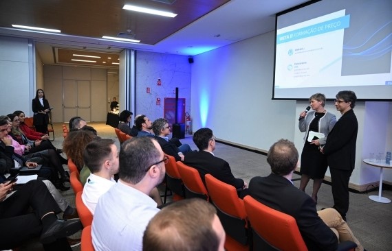 Imagem de um auditório com luzes azuis e cadeiras laranjas. À frente, vemos a vice-presidente da CCEE, Talita Porto, e o diretor-geral da PSR, Luiz Barroso