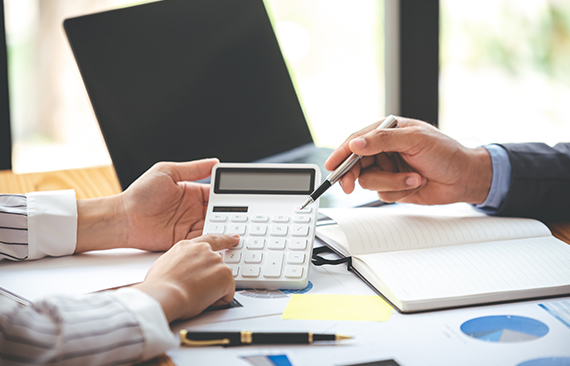 Imagem mostra as mão de dois executivos sobre uma mesa de madeira. Eles usam calculadoras, cadernos e lápis.