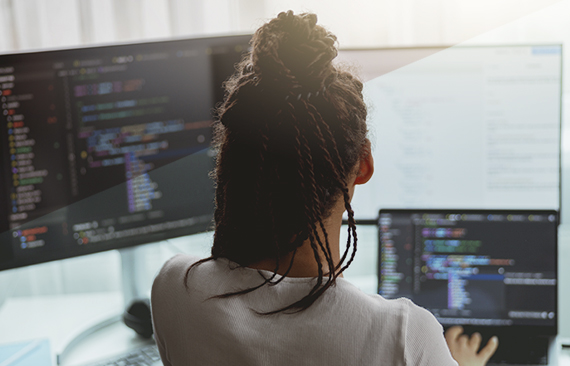 A imagem mostra as costas de uma mulher negra, sentada em frente a telas de computador.