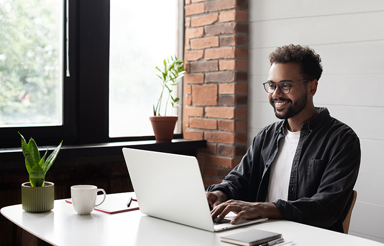 A imagem mostra um homem negro que sorri em frente ao seu computador