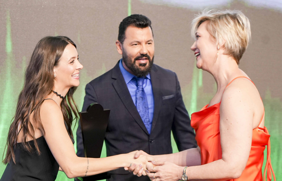 Talita Porto usando vestido vermelho cumprimenta uma mulher sendo observada por um homem de barba