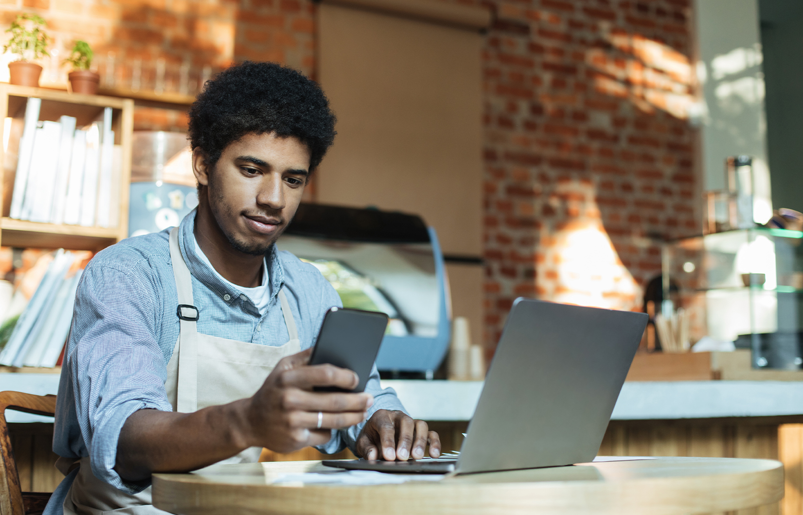 Imagem mostra um empresário em seu negócio. Ele usa um notebook e olha o seu celular.