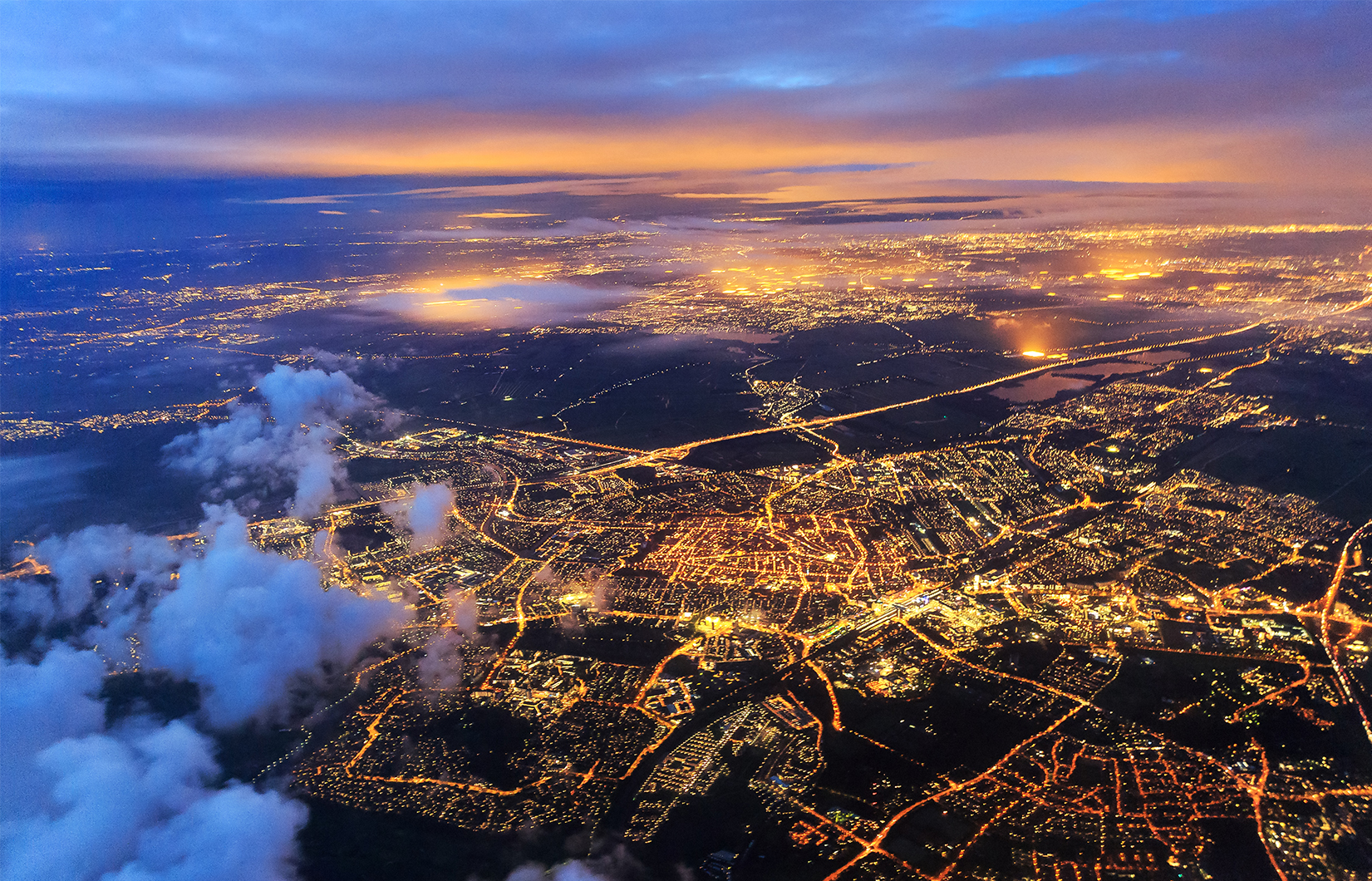 Imagem aérea de uma cidade ao entardecer, com as luzes acesas de vias e construções