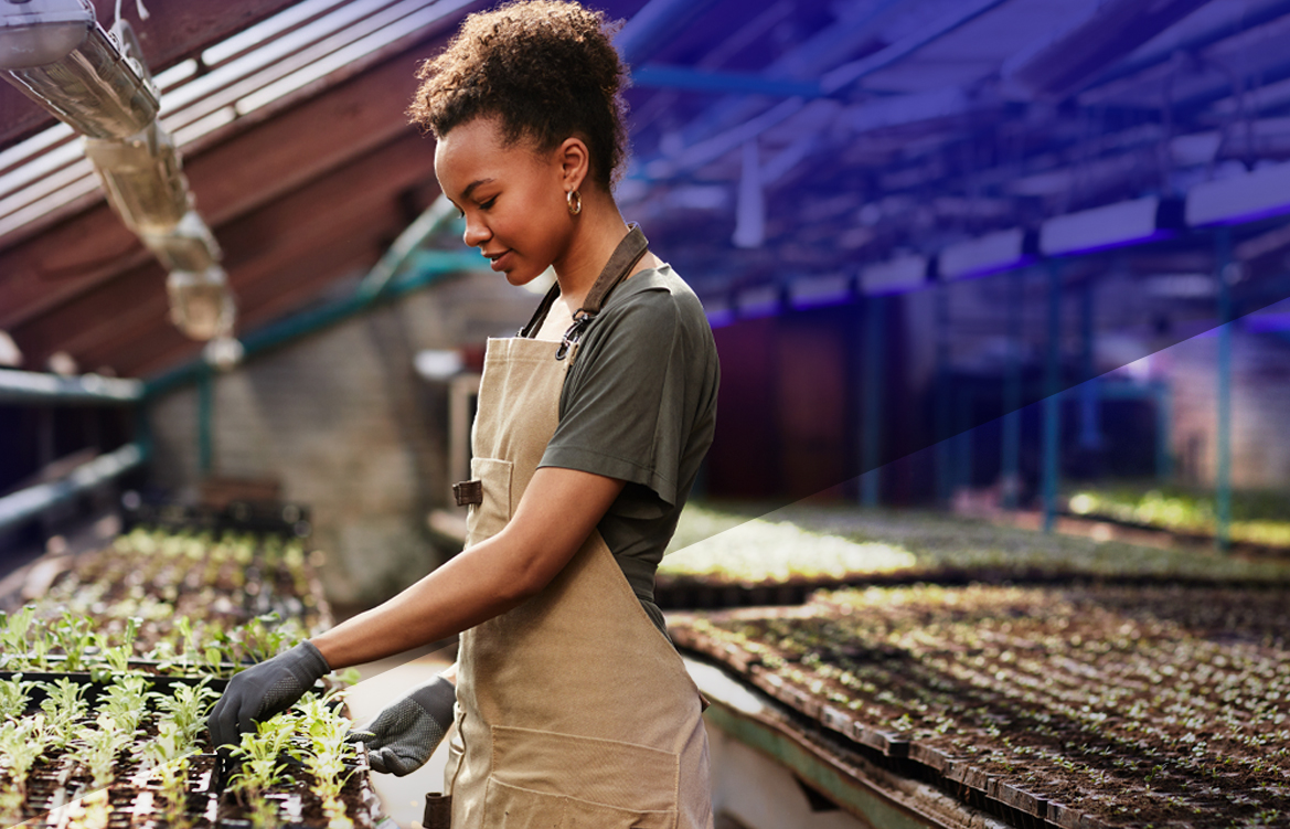 A imagem mostra uma mulher negra que trabalha em um hortifruti. Ela veste um macacão azul e sorri enquanto manuseia verduras.