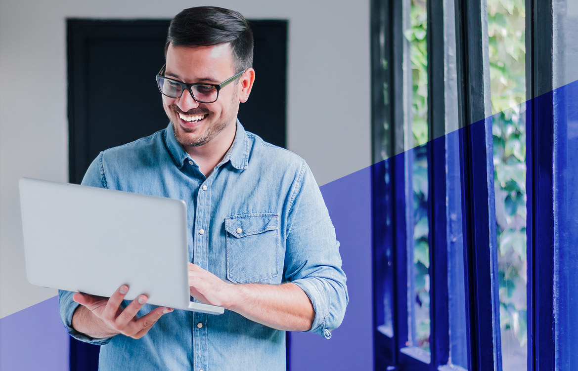 A imagem mostra um homem de camisa azul, em pé, que segura seu notebook com as mãos. Ele olha para a tela do computador e sorri.